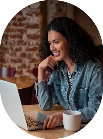 A woman with black skin and curly hair, wearing a black and white striped shirt with a denim jacket over it, smiling while working on her laptop with a mug of coffee next to her.